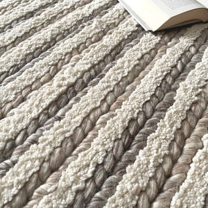 May include: Close-up of a textured rug with alternating stripes of cream and various shades of gray. The rug has a braided design with a soft, plush texture. An open book rests on the rug, suggesting a cozy reading nook.