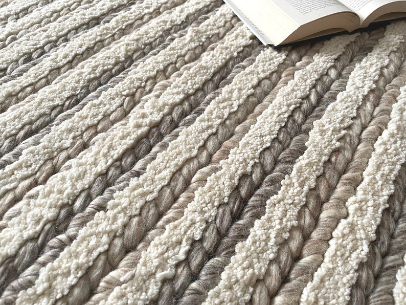 May include: Close-up of a textured rug with alternating stripes of cream and various shades of gray. The rug has a braided design with a soft, plush texture. An open book rests on the rug, suggesting a cozy reading nook.