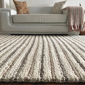 May include: A textured, neutral-toned area rug with vertical stripes of cream, light brown, and gray. The rug is placed on a wooden floor in front of a white sofa. The rug's design adds a cozy and inviting feel to the living space.