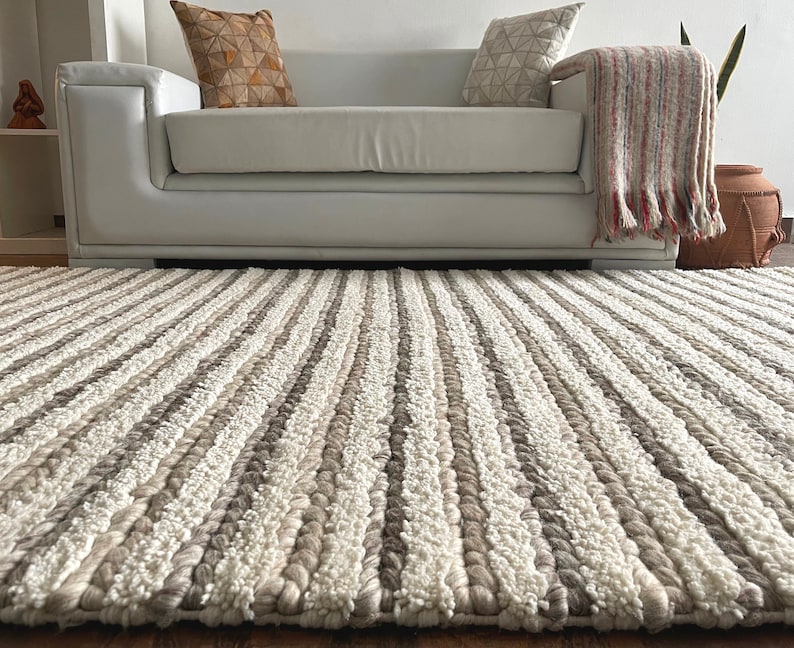 May include: A textured, neutral-toned area rug with vertical stripes of cream, light brown, and gray. The rug is placed on a wooden floor in front of a white sofa. The rug's design adds a cozy and inviting feel to the living space.