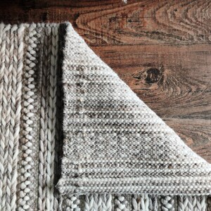 May include: Close-up of a woven rug with a textured, braided design. The rug is a light gray color with hints of brown. The rug is folded over, revealing the underside of the weave.