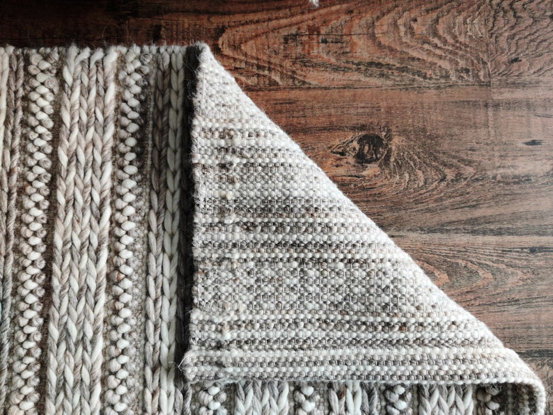 May include: Close-up of a woven rug with a textured, braided design. The rug is a light gray color with hints of brown. The rug is folded over, revealing the underside of the weave.