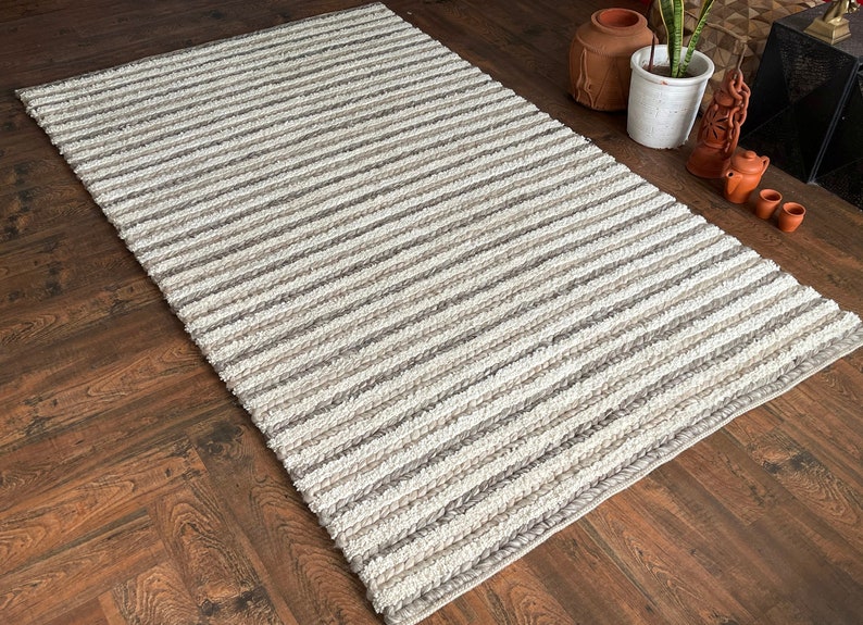 May include: A rectangular rug with a striped pattern in shades of white, gray, and beige. The rug is made of a woven material and is placed on a dark wood floor. The rug's design features alternating stripes of varying widths.