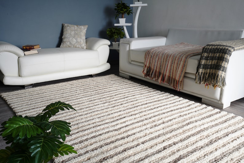 May include: A living room scene featuring a cream-colored, striped rug. Two white leather sofas are positioned on either side of the rug. Throw blankets in neutral tones and a decorative pillow add warmth to the space. A potted plant is in the foreground.