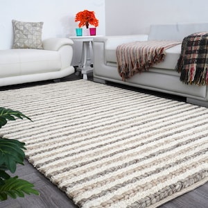 May include: A large, rectangular rug with a striped pattern in shades of cream, beige, and gray. The rug is placed on a dark wood floor in a living room setting, with white sofas and decorative pillows visible in the background.