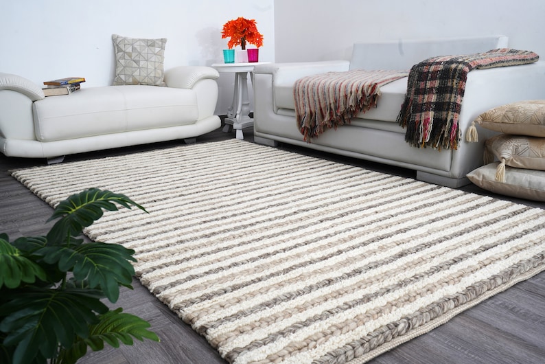 May include: A large, rectangular rug with a striped pattern in shades of cream, beige, and gray. The rug is placed on a dark wood floor in a living room setting, with white sofas and decorative pillows visible in the background.