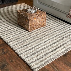May include: A rectangular, striped area rug in shades of cream, gray, and brown. The rug is placed on a dark wood floor. A square, brown leather ottoman with a book on top sits on the rug. A white sofa is in the background.