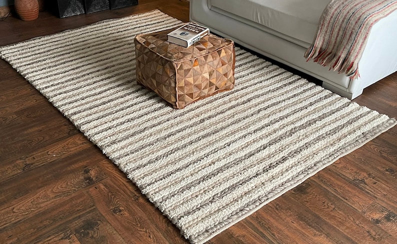 May include: A rectangular, striped area rug in shades of cream, gray, and brown. The rug is placed on a dark wood floor. A square, brown leather ottoman with a book on top sits on the rug. A white sofa is in the background.