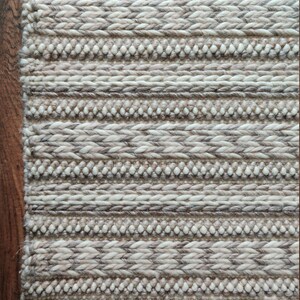 May include: A close-up of a woven rug with a cream and brown striped pattern. The rug has a textured surface and is made of natural fibers.