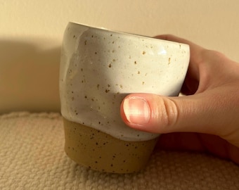 May include: A white ceramic mug with brown speckles and a brown rim. The mug is speckled with brown flecks and has a light brown glaze on the bottom.