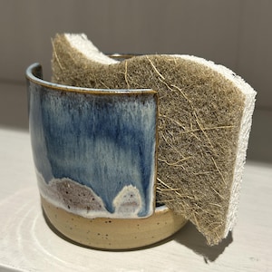 May include: A ceramic sponge holder with a blue and white glazed upper section and a speckled beige base. The holder features a wide U-shaped cutout on the front and contains a rectangular, dual-textured scrubbing sponge with a brown fibrous side.