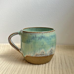 May include: A handcrafted ceramic mug with a rounded form and a robust handle. The mug displays a colour gradient, shifting from light green at the top to a brown base. The interior is a solid green colour.