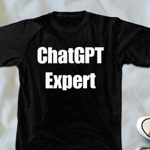 May include: Black t-shirt with the text "ChatGPT Expert" in white. The shirt is displayed with a pair of blue jeans and tan trainers with white laces and soles.
