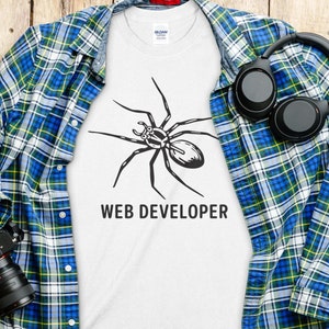 May include: White t-shirt with a black spider graphic and the text "WEB DEVELOPER". The shirt is laid on a blue, green, and yellow plaid shirt. A black camera and headphones are also visible.