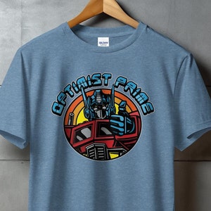 Puede incluir: Una camiseta azul con un gráfico de Optimus Prime, un robot de la franquicia Transformers, en su forma de camión. El gráfico está en un círculo con el texto "Optimist Prime" alrededor.