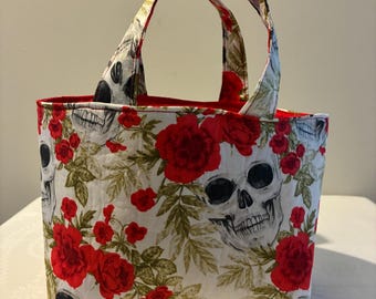 Puede incluir: Un bolso de tela acolchada con un patrón repetitivo de calaveras humanas, rosas rojas vibrantes y hojas verdes sobre un fondo blanco. El bolso tiene dos asas de tela a juego y un forro interior de color rojo sólido visible en la abertura.