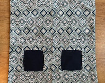 Puede incluir: Un artículo de tela estampada con dos bolsillos cuadrados negros. La tela tiene un diseño geométrico repetido en tonos verdes, azules y beige. Las esquinas superiores tienen clips metálicos dorados.