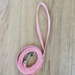Puede incluir: Una correa de perro rosa con un clip plateado. La correa est&aacute; enrollada sobre una superficie de madera.