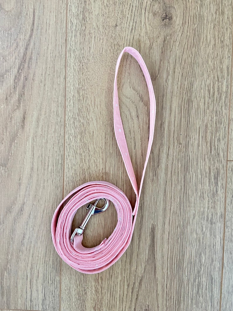 Puede incluir: Una correa de perro rosa con un clip plateado. La correa est&aacute; enrollada sobre una superficie de madera.
