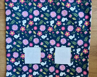 Puede incluir: Una bolsa de tela cuadrada con fondo azul oscuro y un patrón repetitivo de conejos blancos, flores rosas y hojas verdes. La bolsa incluye dos aberturas cuadradas blancas y dos clips giratorios de metal sujetos en las esquinas superiores.