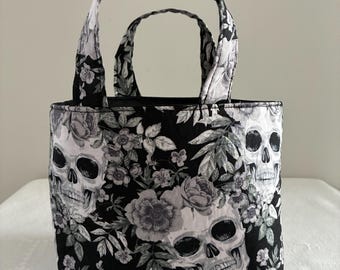Puede incluir: Un bolso de tela con un patrón en blanco y negro de calaveras humanas intercaladas con diversos elementos florales. El bolso tiene dos asas de tela a juego y una estructura rectangular con base plana, mostrando un diseño botánico repetitivo.