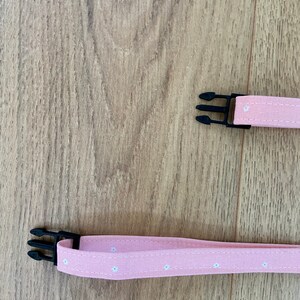 Puede incluir: Dos correas de tela rosa con patrones de flores blancas y cierres de pl&aacute;stico negro. Las correas son para un collar o arn&eacute;s para perros.