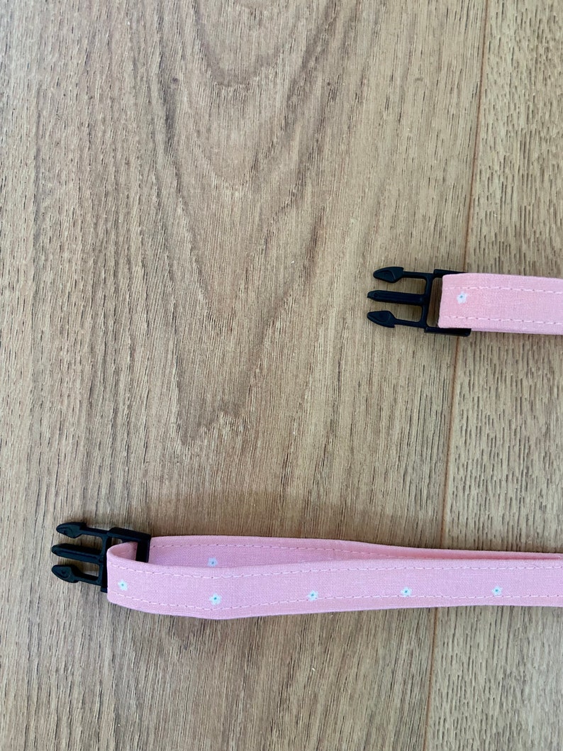 Puede incluir: Dos correas de tela rosa con patrones de flores blancas y cierres de pl&aacute;stico negro. Las correas son para un collar o arn&eacute;s para perros.