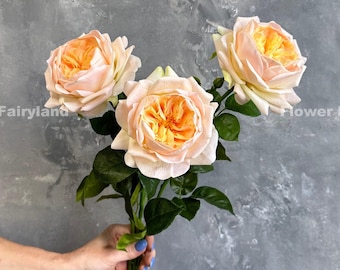 May include: A bouquet of three peach-colored artificial roses with yellow centers. The roses are arranged in a hand holding the stems.
