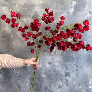 Puede incluir: Un ramo de flores rojas artificiales con un diseño único en forma de campana. Las flores se sostienen en una mano sobre un fondo gris. Los tallos son verdes y las flores son de un rojo intenso y rico. El texto "Flower Fair and Chicago" es visible.