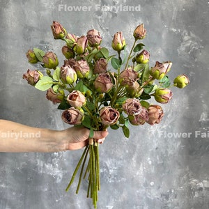 Op de afbeelding: Een boeket van kunstmatige, stoffige rozen in roze tinten met groene stelen en bladeren. De rozen zijn gerangschikt in een hand die de stelen vasthoudt.