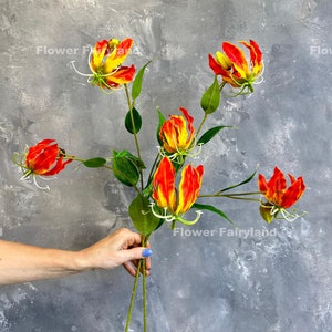 Può includere: Un bouquet di fiori artificiali dai vivaci petali rossi e gialli, con steli e foglie verdi. I fiori sono tenuti da una persona. Lo sfondo è un muro grigio strutturato. Il testo "Flower Fairyland" è visibile.