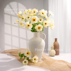 Peut inclure: Un bouquet de fleurs artificielles de couleur cr&egrave;me dans un vase en c&eacute;ramique blanche. Le vase a une large ouverture et une poign&eacute;e. Les fleurs sont dispos&eacute;es de mani&egrave;re l&acirc;che et naturelle. Le vase est pos&eacute; sur une nappe de table beige.