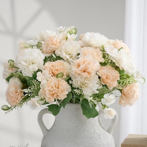 May include: A full bouquet of artificial carnations in a textured, light gray vase. The flowers are a mix of cream and peach colors, with green foliage. The vase has two handles and a wide opening, perfect for a floral arrangement.