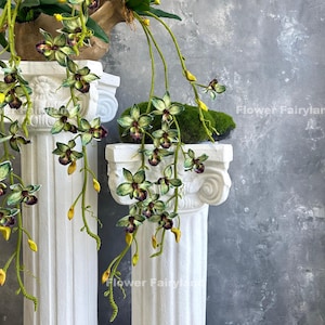 May include: Two white columns with green and purple orchid flowers hanging from them. The orchids are cascading down the columns and the background is a grey textured wall.