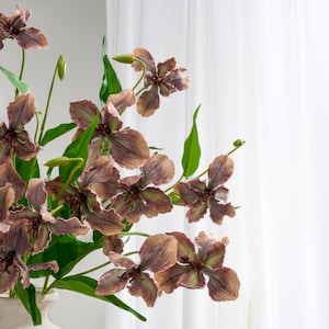 May include: Close-up of a floral arrangement featuring artificial orchids. The orchids have brown and pink petals with green accents and stems. The arrangement includes green leaves and is set against a white background.