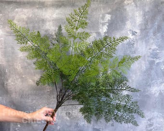 May include: A bright green artificial fern stem with multiple fronds. The fern has a dark brown stem and is held against a grey background. The fern's leaves are detailed and create a full, textured appearance. Suitable for floral arrangements and home decor.