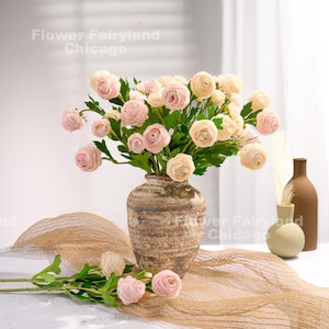 Peut inclure: Un bouquet de fleurs de renoncule en soie rose p&acirc;le dans un vase en c&eacute;ramique brun. Le vase a une surface textur&eacute;e et repose sur une surface textur&eacute;e beige. Les fleurs sont dispos&eacute;es dans un style l&acirc;che et naturel.