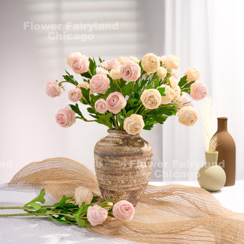 Peut inclure: Un bouquet de fleurs de renoncule en soie rose p&acirc;le dans un vase en c&eacute;ramique brun. Le vase a une surface textur&eacute;e et repose sur une surface textur&eacute;e beige. Les fleurs sont dispos&eacute;es dans un style l&acirc;che et naturel.