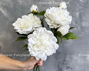 May include: A bouquet of large, white peony flowers with green leaves and stems. The flowers are fully bloomed, showcasing their layered petals. The background is a textured gray surface. The text "Flower Fairyland" is visible.