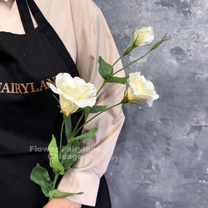 May include: A stem of artificial cream-colored lisianthus flowers with ruffled petals, green leaves, and a closed bud. The stem is held against a black apron featuring the gold text "FAIRYLAND" and "Flower Fairyland Chicago".