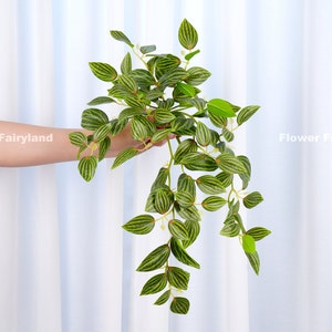 May include: A long, trailing artificial plant with green leaves that have a striped pattern. The leaves are arranged in a cascading pattern, giving the plant a flowing appearance.
