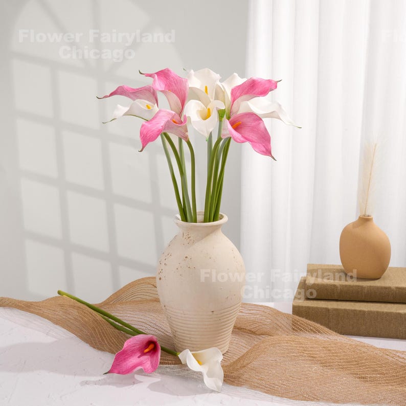 May include: A bouquet of artificial calla lilies in a cream-colored vase. The lilies have white and pink petals with yellow centers. The vase sits on a textured surface with a small, tan vase and books in the background. The text "Flower Fairyland Chicago" is visible.