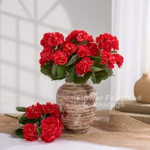 Puede incluir: Un ramo de flores rojas vibrantes en un jarrón marrón texturizado. El arreglo incluye hojas verdes exuberantes y flores rojas adicionales sobre una superficie de arpillera. Un jarrón más pequeño, de color beige, es visible en el fondo.