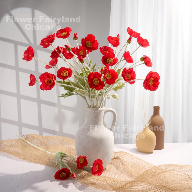 May include: A bouquet of red poppy flowers in a white ceramic vase. The vase has a handle and a textured surface. The flowers are arranged in a vase on a brown woven surface.