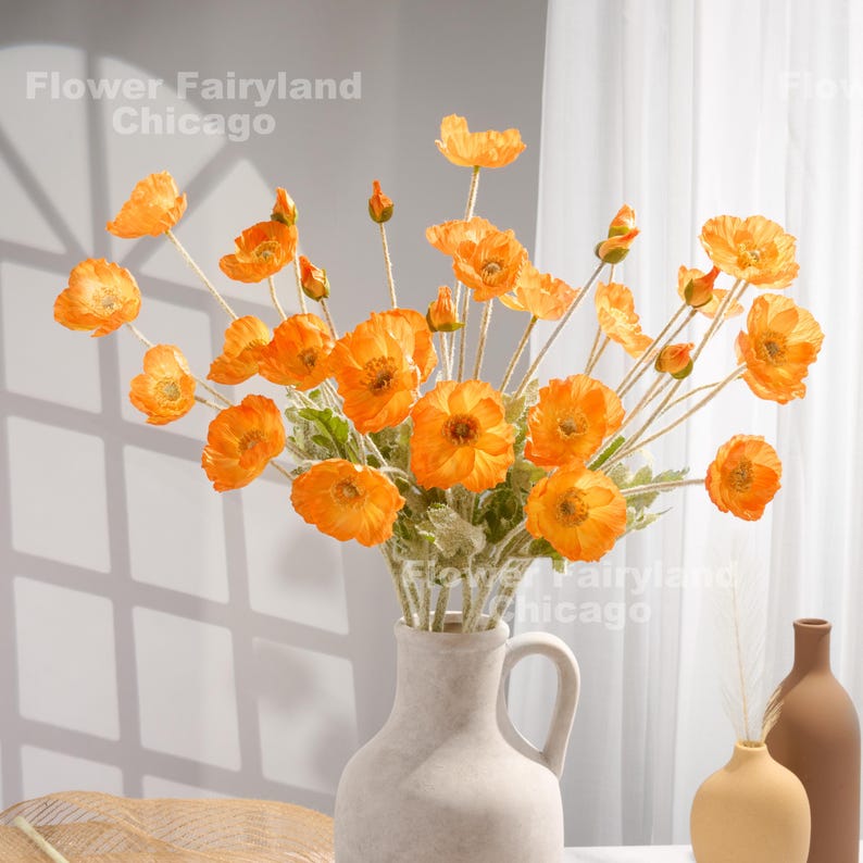 May include: A bouquet of orange silk poppy flowers in a white ceramic vase. The flowers are arranged in a vase with a handle. The vase is sitting on a wooden surface.