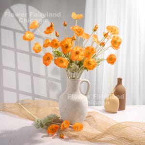 May include: A bouquet of orange silk poppy flowers in a white ceramic vase. The vase has a handle and a round base. The flowers are arranged in a vase on a brown woven surface.