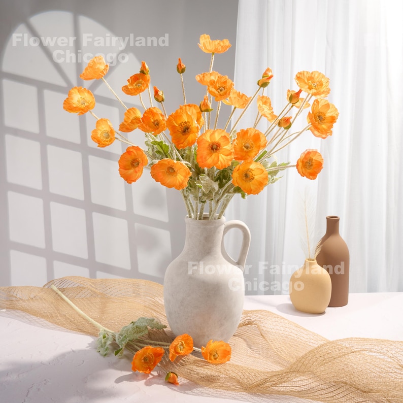 May include: A bouquet of orange silk poppy flowers in a white ceramic vase. The vase has a handle and a round base. The flowers are arranged in a vase on a brown woven surface.