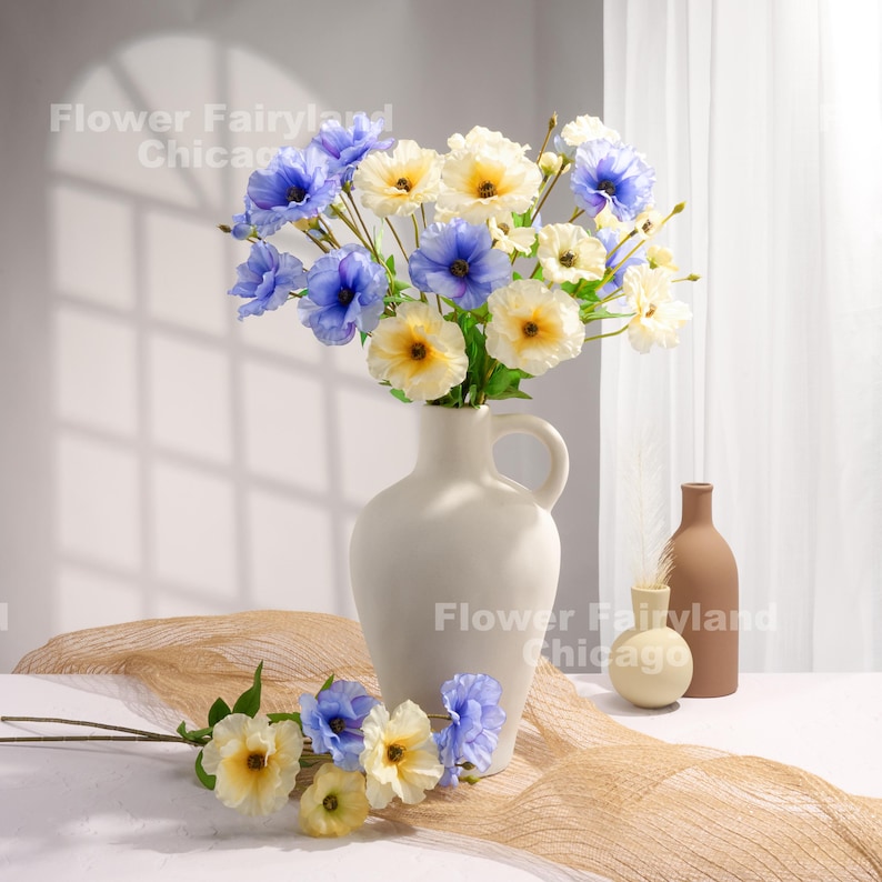 Peut inclure: Un bouquet de fleurs artificielles bleues et jaunes dans un vase en c&eacute;ramique blanc. Le vase a une anse et un corps arrondi. Les fleurs sont dispos&eacute;es dans un style l&acirc;che et naturel. Les fleurs sont pos&eacute;es sur une table avec une nappe en tissu beige.