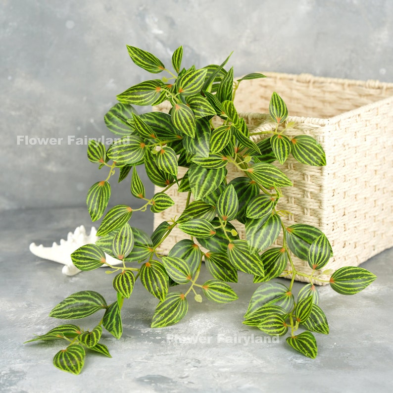 May include: Artificial green plant with striped leaves hanging over a woven basket. The plant has a vine-like appearance and is ideal for adding greenery to a home or office.