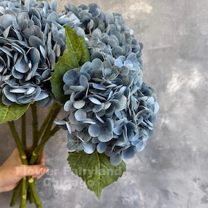 Könnte beinhalten: Ein Strauß blauer Hortensienblüten mit grünen Blättern. Die Blüten haben mehrere Schichten von Blütenblättern und sind staubblau. Die Stiele sind grün. Der Text "Flower Fairyland Chicago" ist sichtbar.